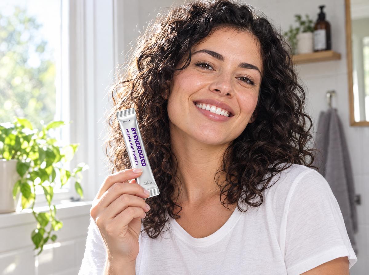 Woman smiling by a sunny bathroom window holding a Byenergized purple mouthwash sachet